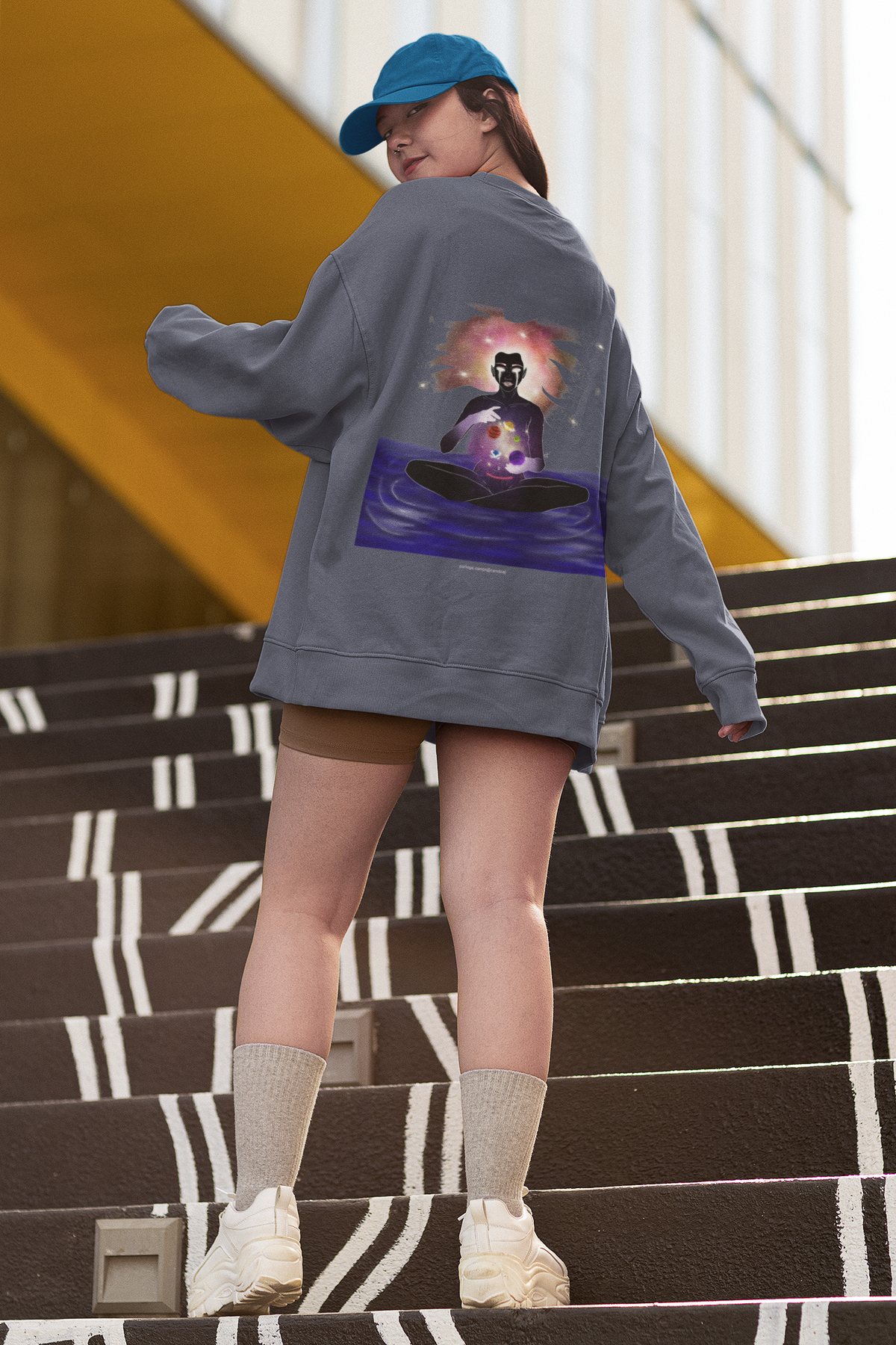Orbit Keeper Garment-Dyed Crewneck in Denim, worn by a woman seen from behind on outdoor stairs, showing the large print on the back. Digital product mockup.