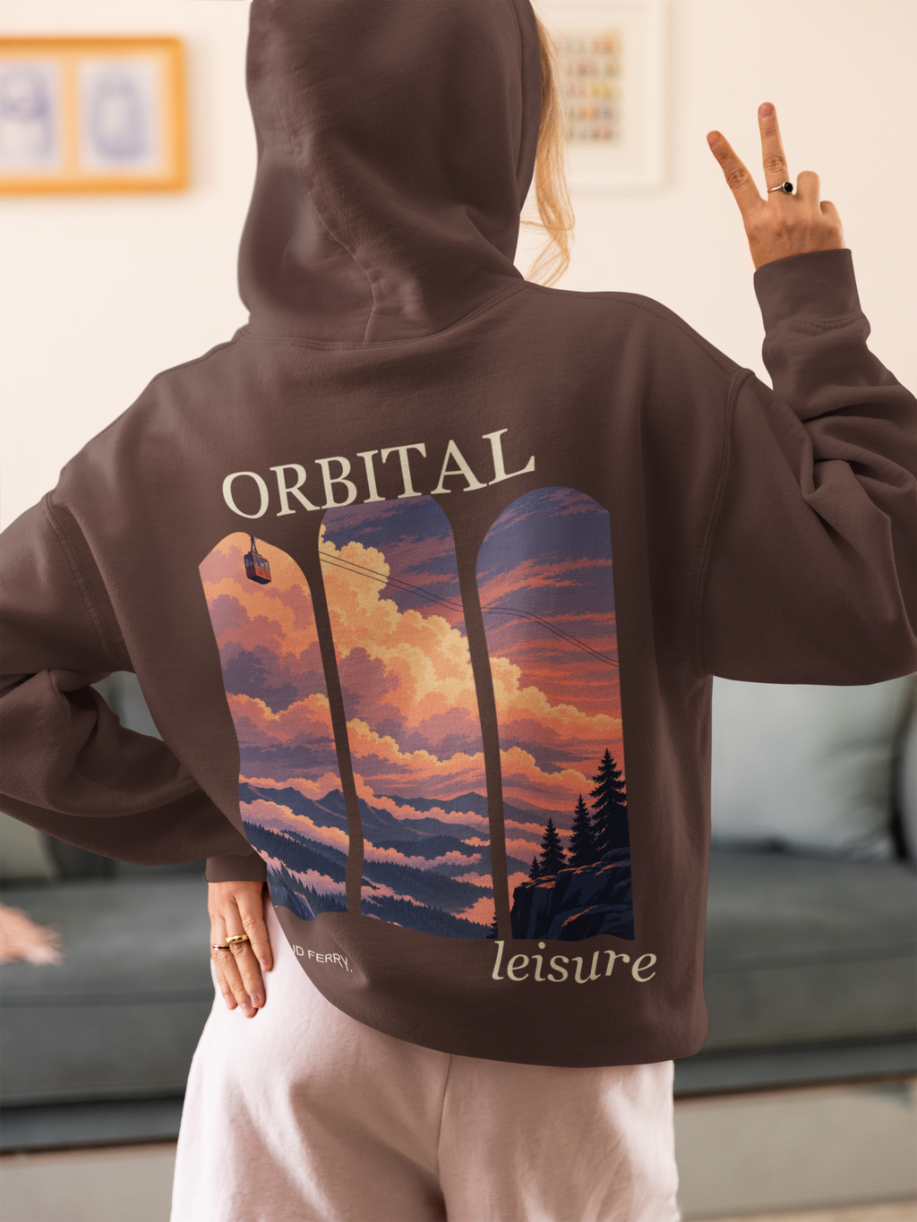 Cloud Ferry Heavyweight Hoodie in Brown, worn by a woman seen from behind at home, making a peace sign, showing the large print on the back. Digital product mockup.