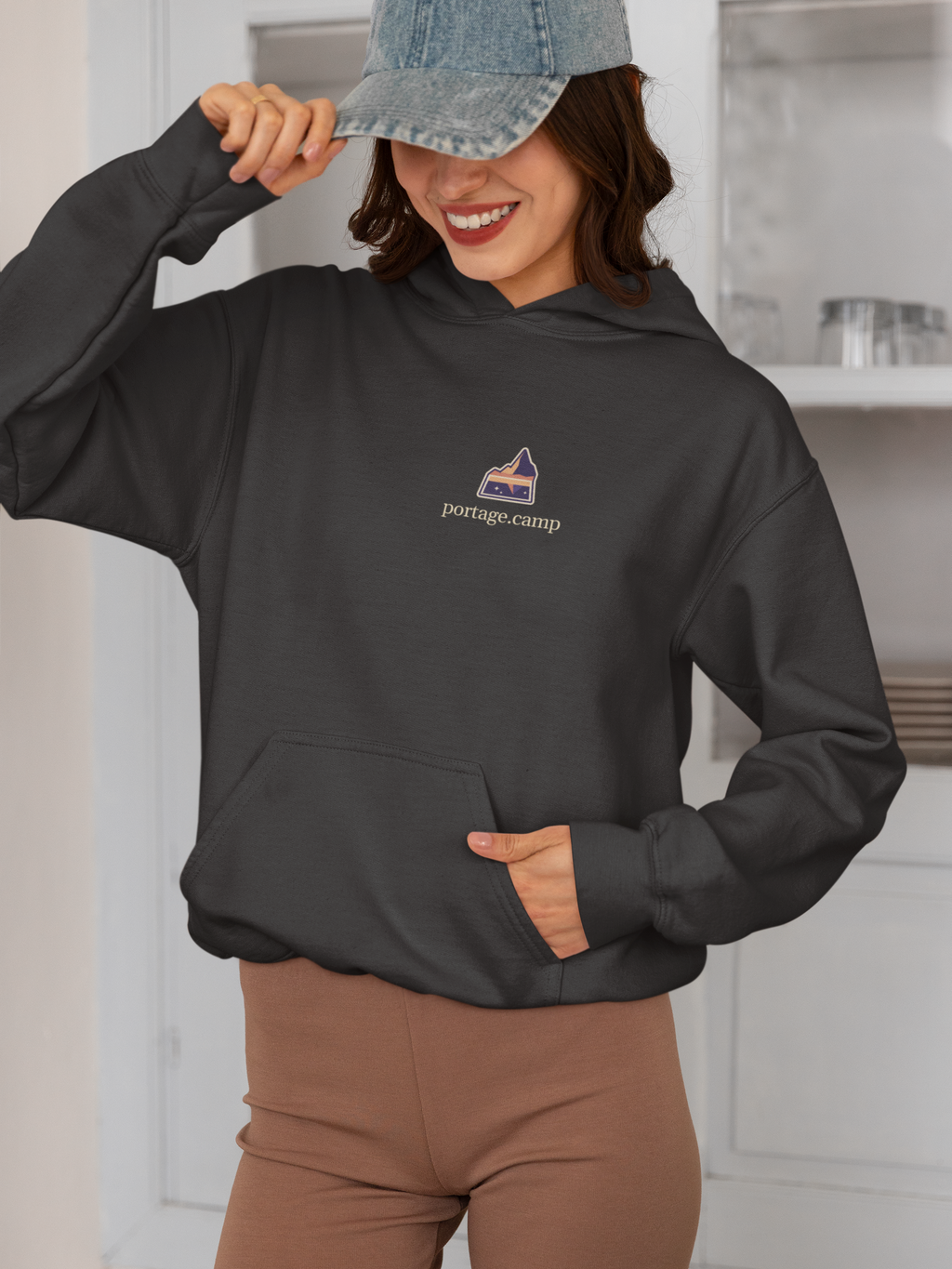 A Different Sky Heavyweight Hoodie in Black, worn by a woman smiling in a kitchen, with the A Different Sky logo and portage.camp on the left chest. Digital product mockup.