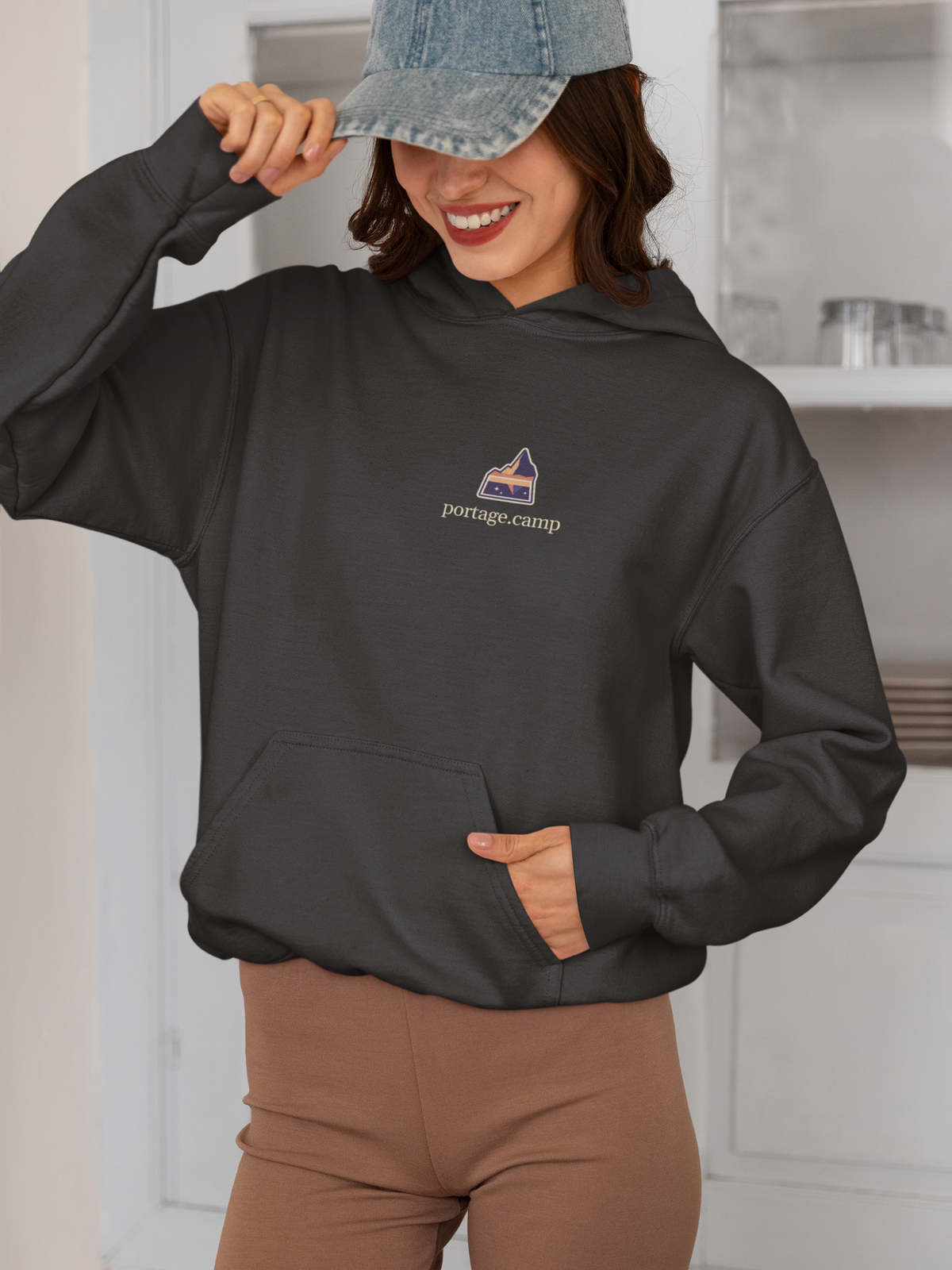 A Different Sky Heavyweight Hoodie in Black, worn by a woman smiling in a kitchen, with the A Different Sky logo and portage.camp on the left chest. Digital product mockup.
