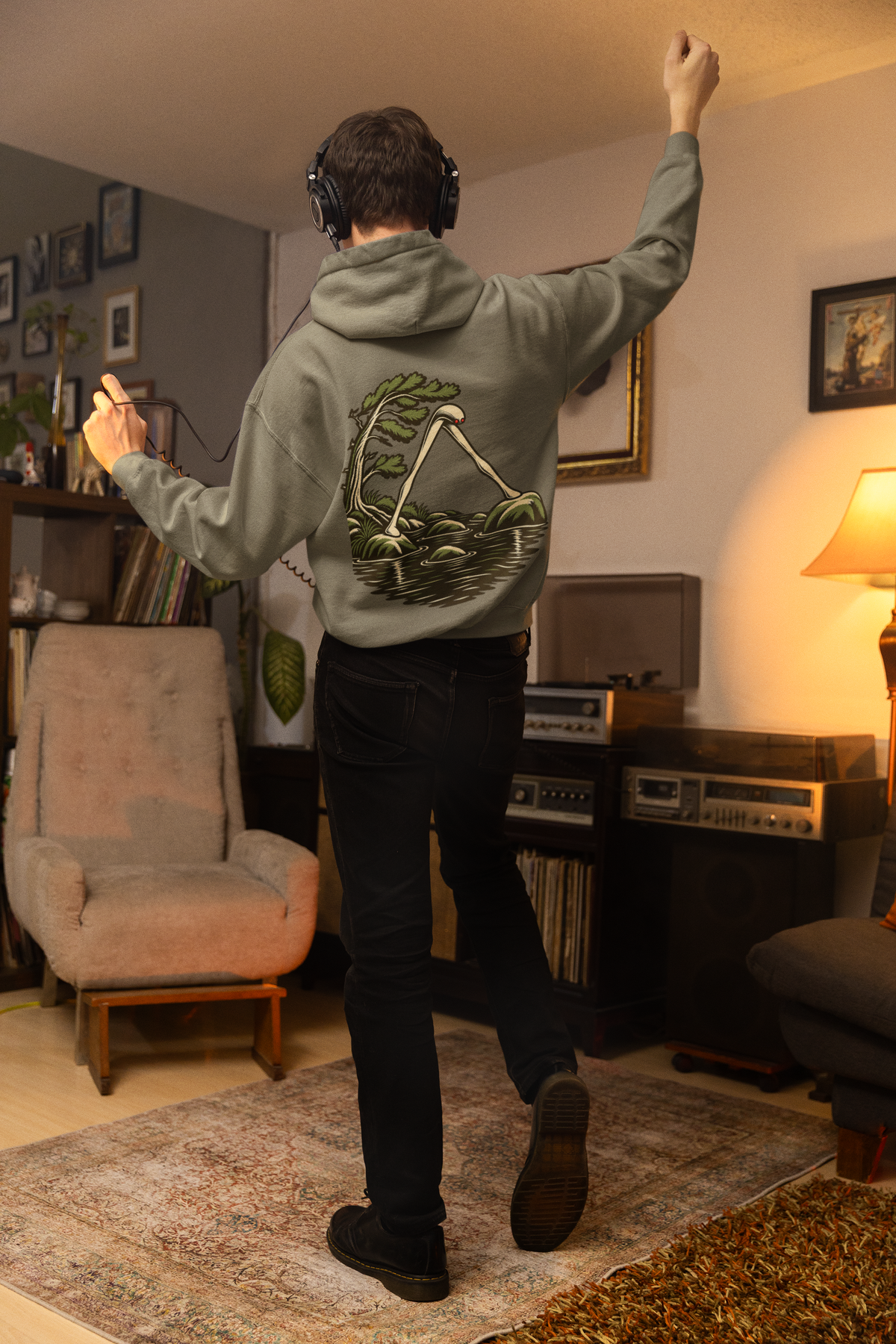 Small Hours Heavyweight Hoodie in Olive, worn by a man seen from behind in a living room at night, showing the large print on the back. Digital product mockup.