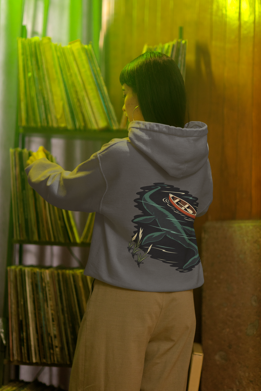 Seen Briefly Midweight Hoodie in Charcoal, worn by a woman seen from behind in a vintage record shop, showing the large print on the back. Digital product mockup.