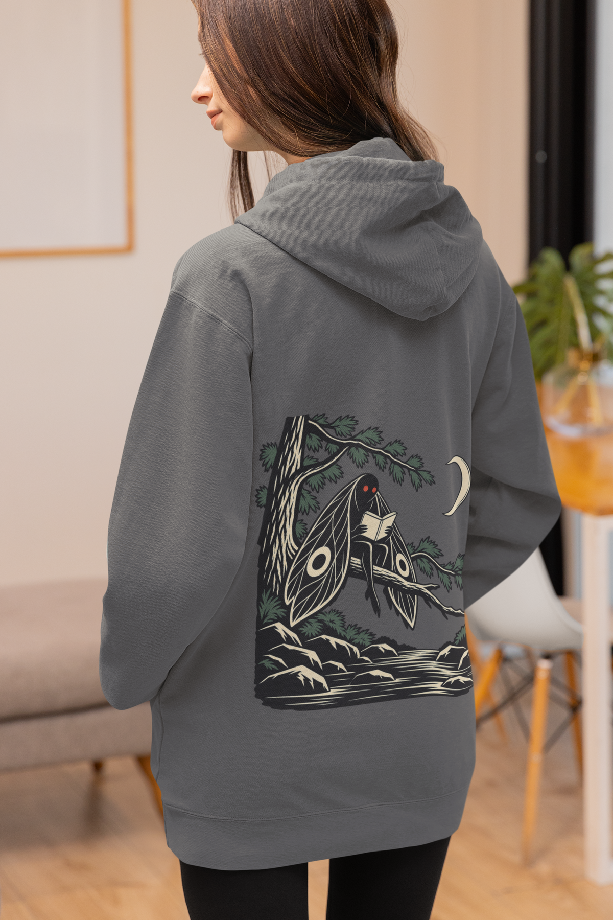Night Library Midweight Hoodie in Charcoal, worn by a woman seen from behind at home, showing the large print on the back. Digital product mockup.
