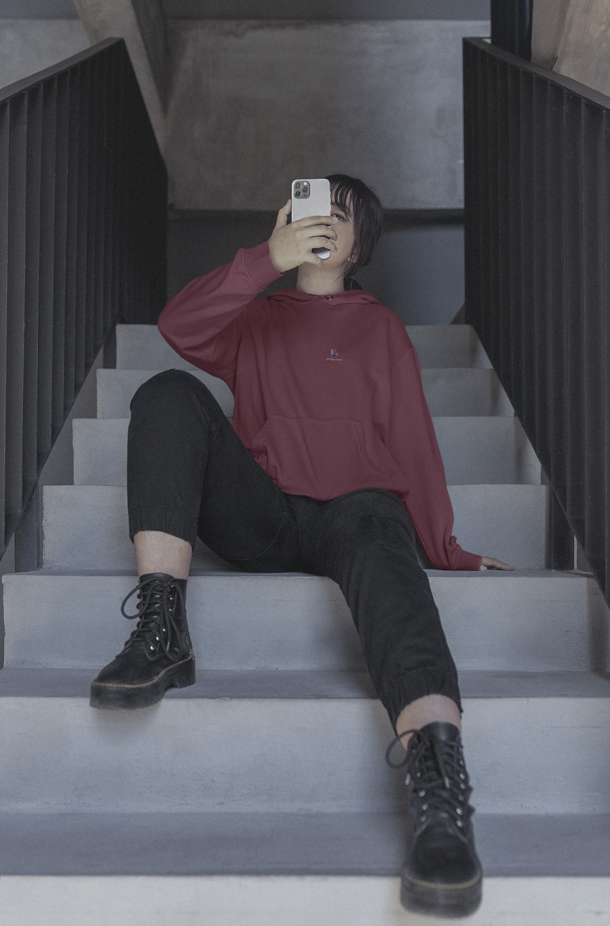 Orbit Keeper Midweight Hoodie in Maroon, worn by a woman seated on a staircase, with the Orbit Keeper logo and portage.camp on the chest. Digital product mockup.