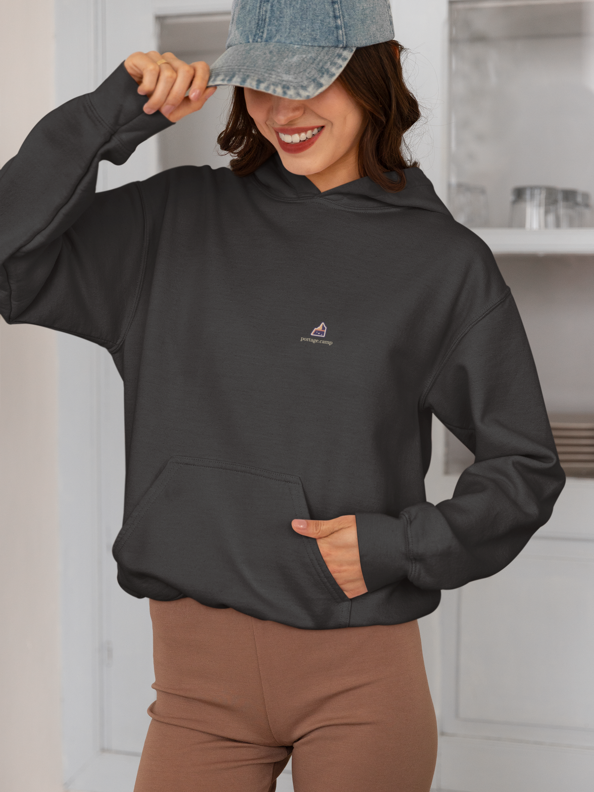 A Different Sky Heavyweight Hoodie in Black, worn by a woman smiling in a kitchen, with the A Different Sky logo and portage.camp on the left chest. Digital product mockup.