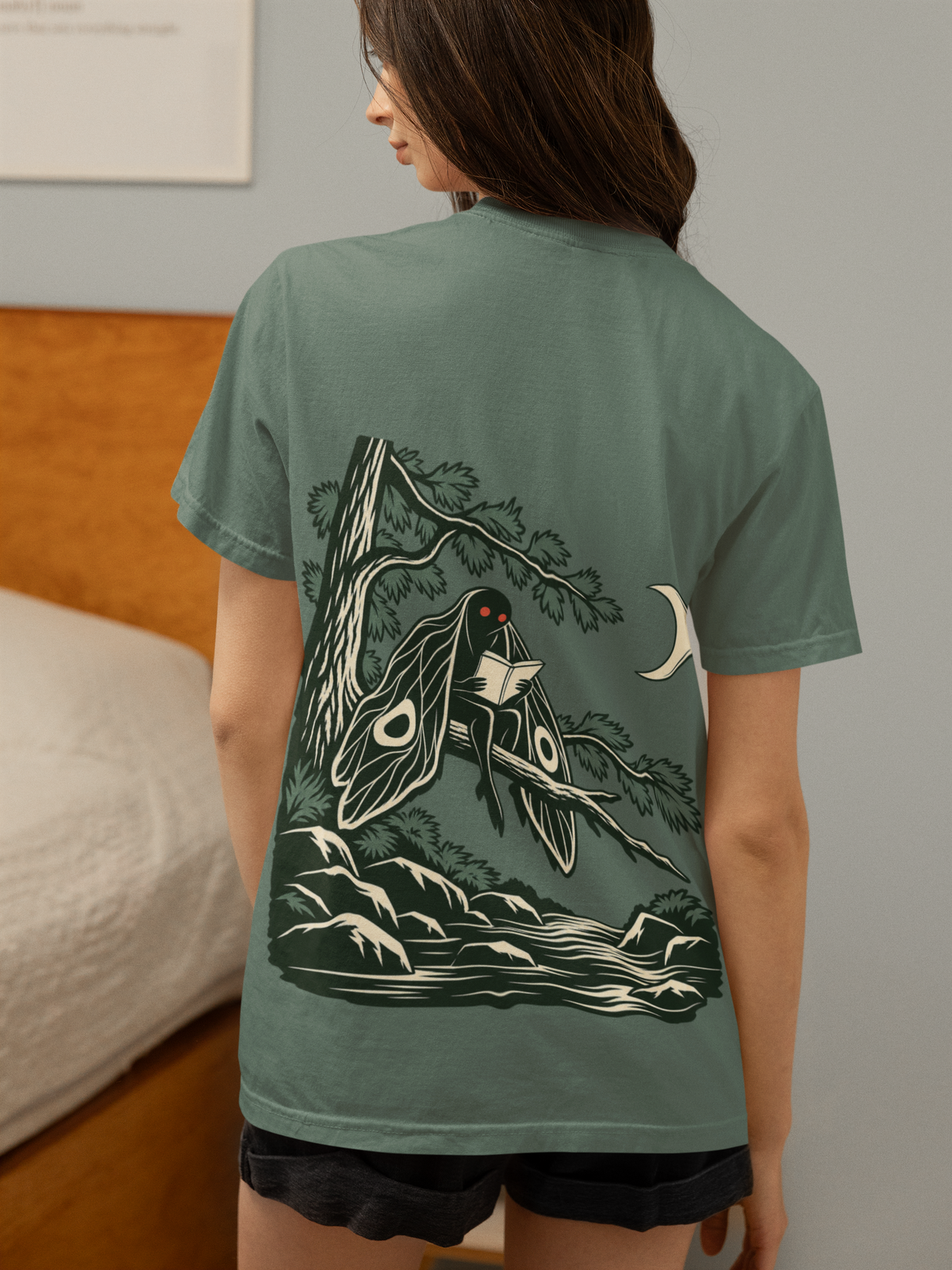 Night Library Garment-Dyed Tee in Blue Spruce, worn by a woman seen from behind in a bedroom at home, showing the large print on the back. Digital product mockup.
