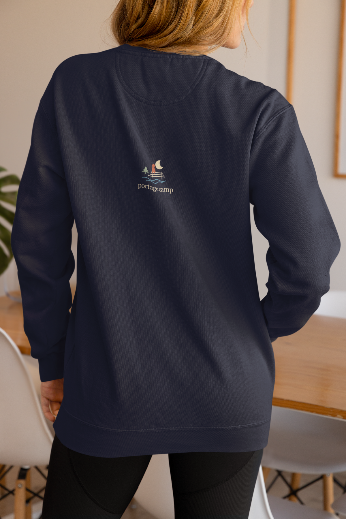 Quiet Hours Garment-Dyed Crewneck (Front Print) in True Navy, worn by a woman seen from behind at home, showing the portage.camp emblem at the upper back. Digital product mockup.