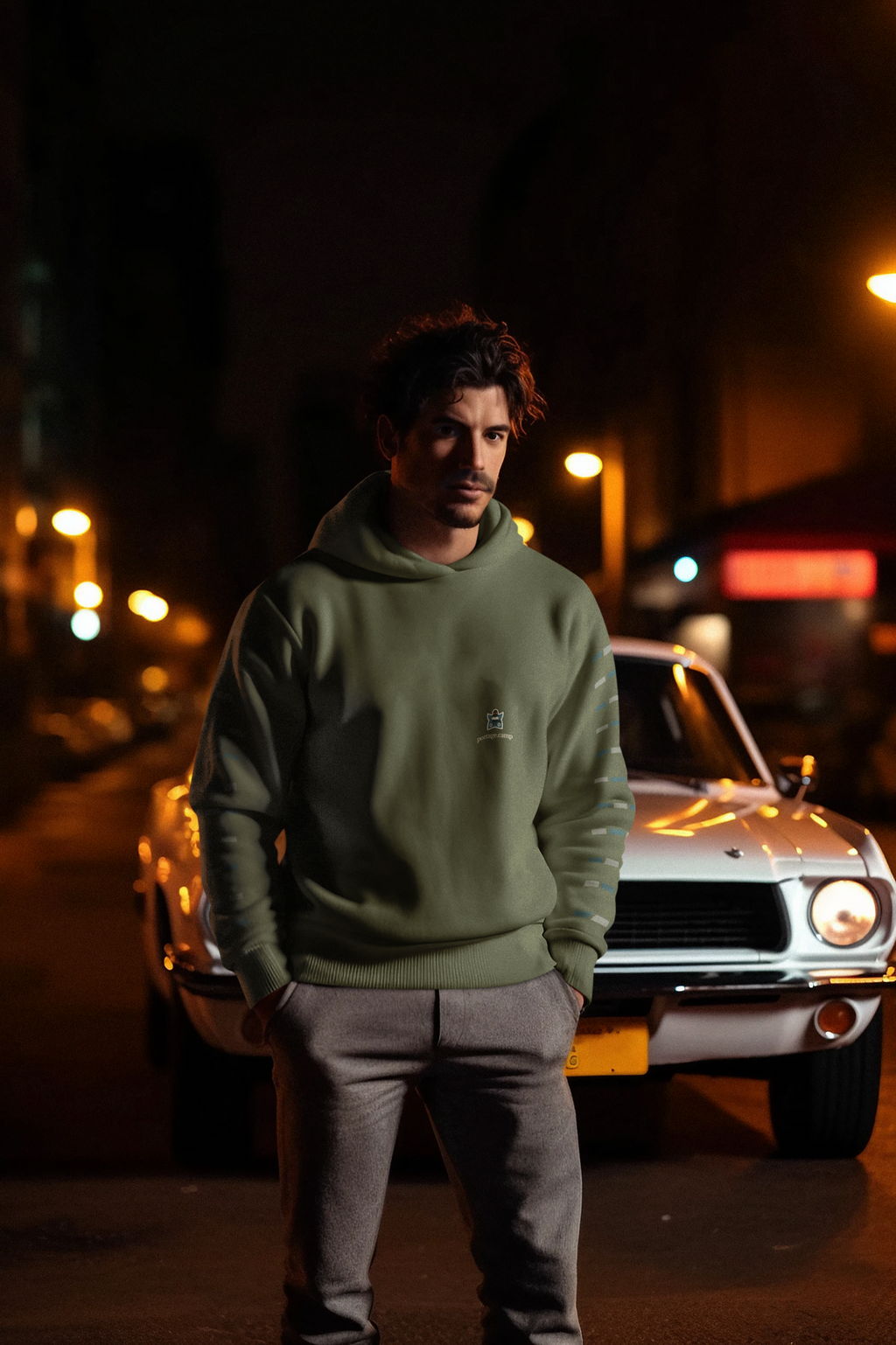 Night Library Midweight Hoodie in Army, worn by a man standing by a classic car at night, with the Night Library logo and portage.camp on the chest. Digital product mockup.
