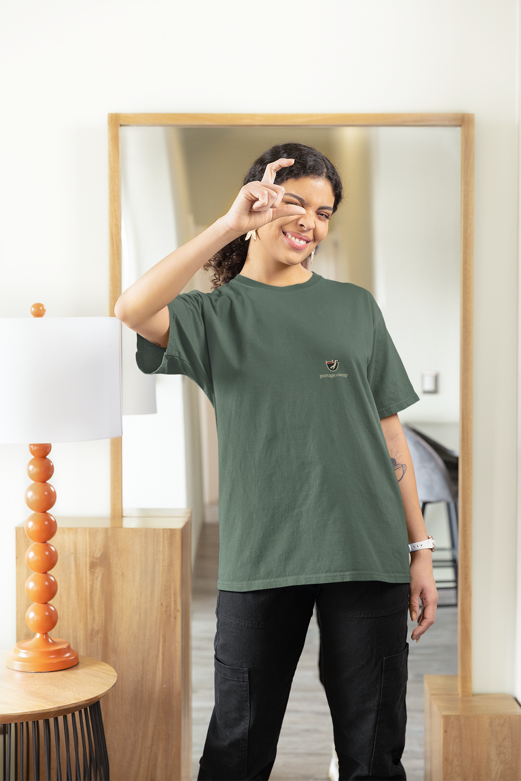 Seen Briefly Garment-Dyed Tee in Blue Spruce, worn by a woman at home, pretending to take a photo, with the Seen Briefly logo and portage.camp on the chest. Digital product mockup.