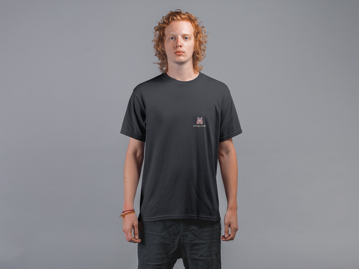Edge of Evening Garment-Dyed Tee in Black, worn by a man against a plain grey wall, with the Edge of Evening logo and portage.camp on the chest. Digital product mockup.