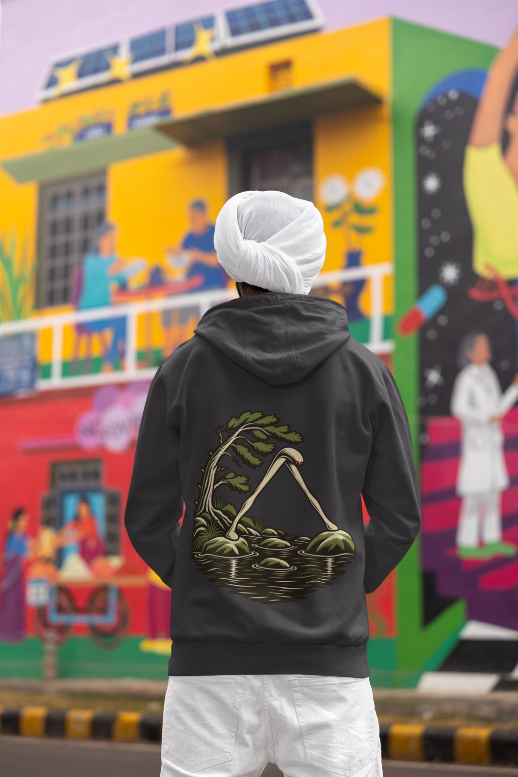 Open Late Heavyweight Hoodie in Black, worn by a man seen from behind in a turban by a painted mural, showing the large print on the back. Digital product mockup.