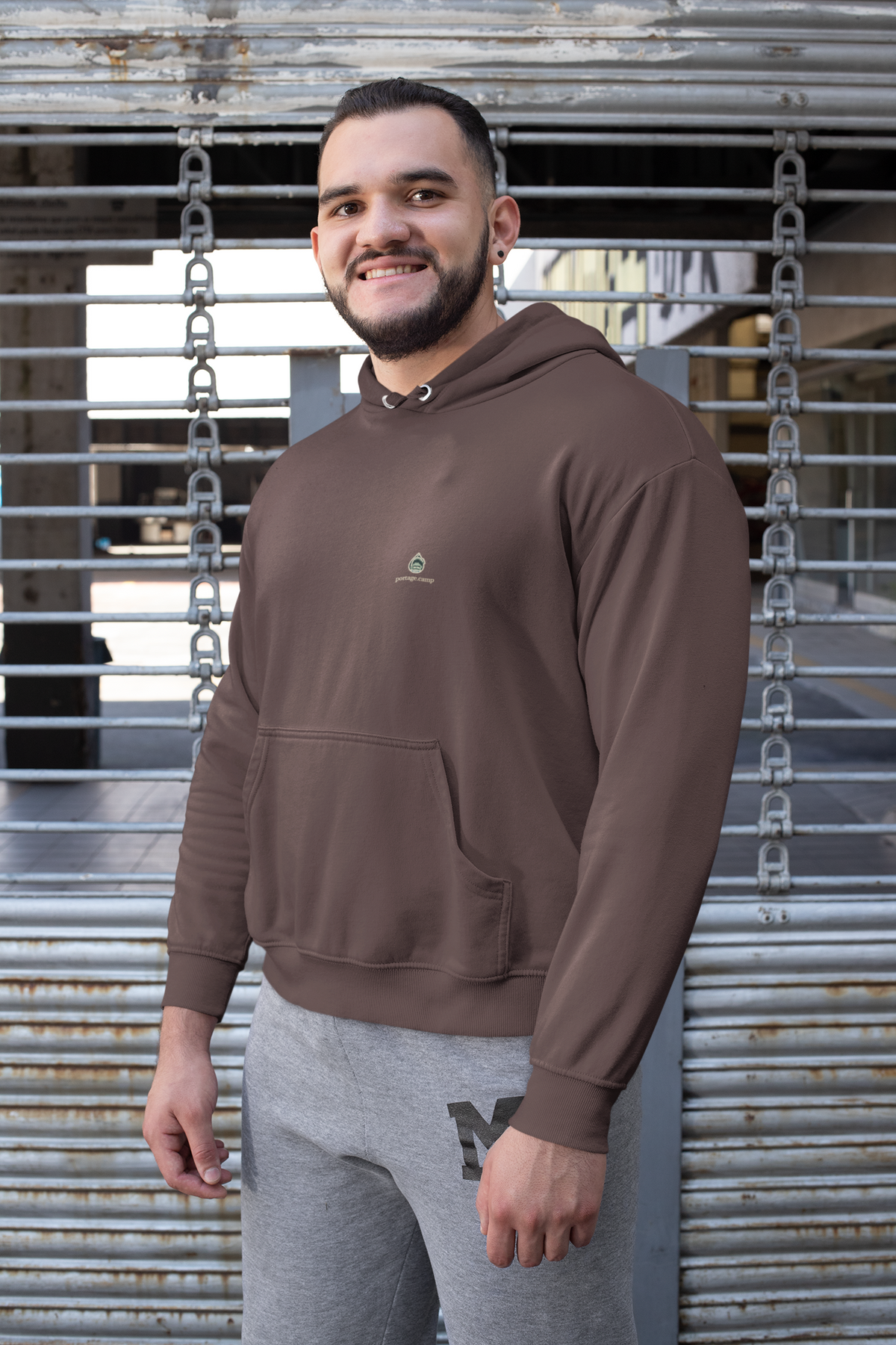 Rain Check Heavyweight Hoodie in Brown, worn by a man against a metal fence, with the Rain Check logo and portage.camp on the left chest. Digital product mockup.
