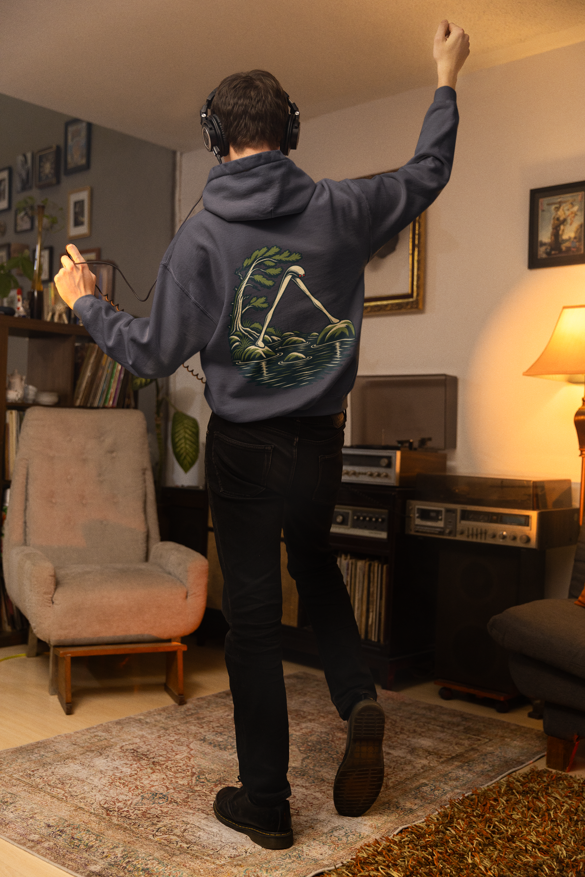 Small Hours Midweight Hoodie in Classic Navy, worn by a man seen from behind in a living room at night, showing the large print on the back. Digital product mockup.