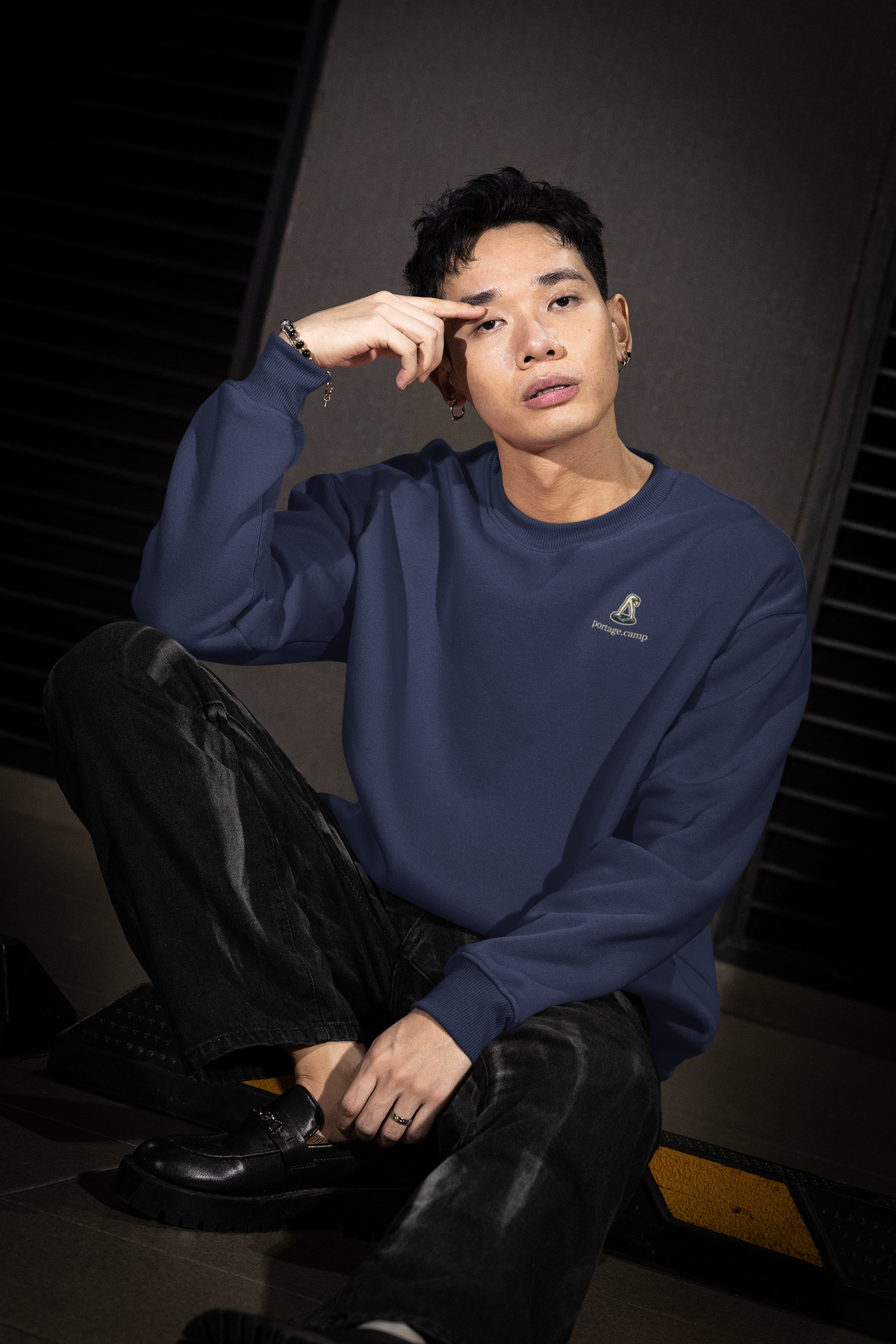 Small Hours Garment-Dyed Crewneck in True Navy, worn by a man seated on a city sidewalk, with the Small Hours logo and portage.camp on the chest. Digital product mockup.
