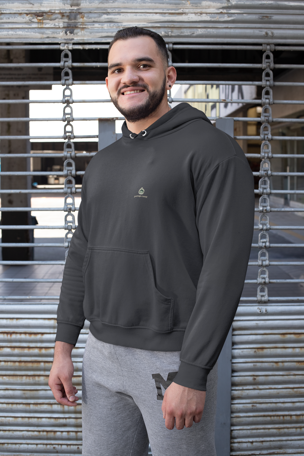 Rain Check Heavyweight Hoodie in Black, worn by a man against a metal fence, with the Rain Check logo and portage.camp on the left chest. Digital product mockup.