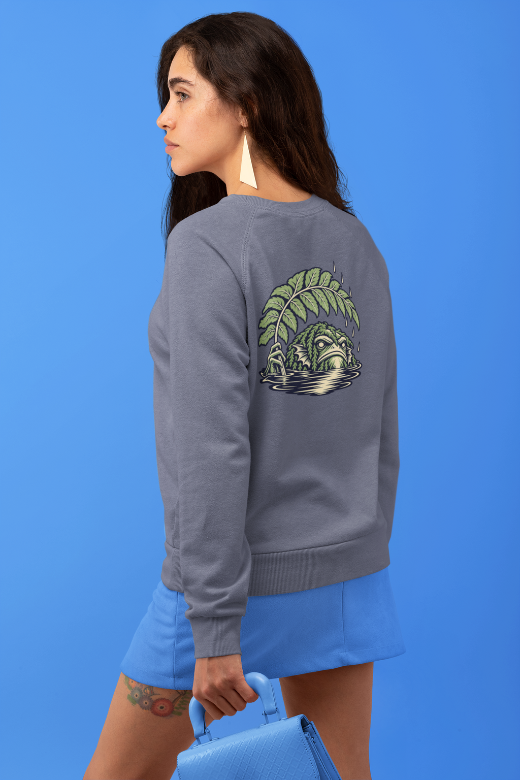 Rain Check Garment-Dyed Crewneck in Denim, worn by a woman seen from behind against a blue backdrop, showing the large print on the back. Digital product mockup.