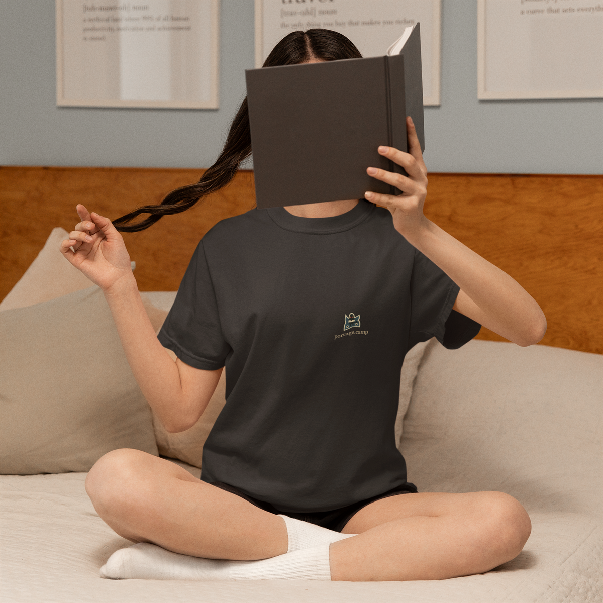 Night Library Garment-Dyed Tee in Black, worn by a woman at home, hiding behind a book, with the Night Library logo and portage.camp on the chest. Digital product mockup.