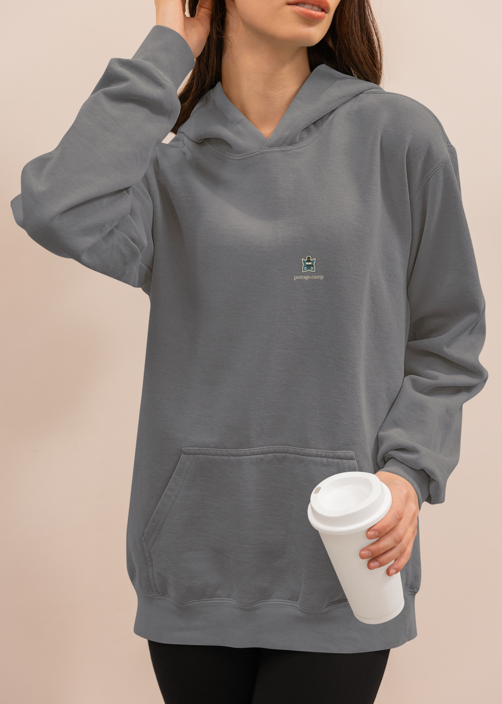 Night Library Midweight Hoodie in Charcoal, worn by a woman at home with a coffee cup, with the Night Library logo and portage.camp on the chest. Digital product mockup.