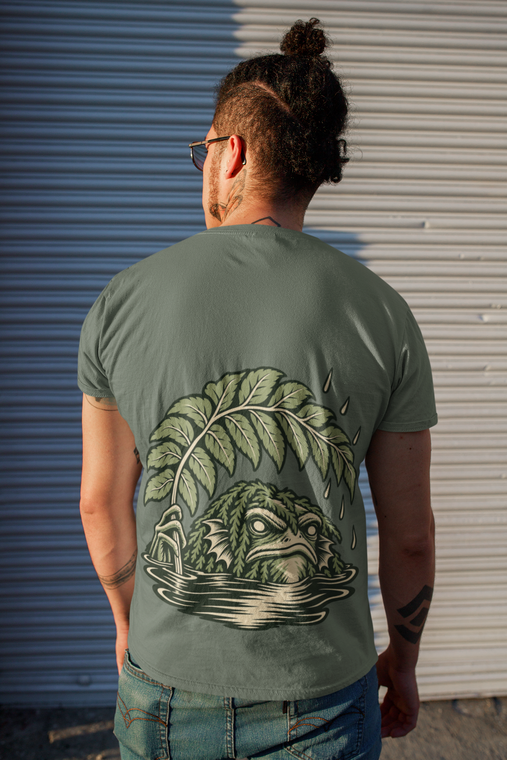 Rain Check Garment-Dyed Tee in Blue Spruce, worn by a man seen from behind on a street beside a metal shutter, showing the large print on the back. Digital product mockup.