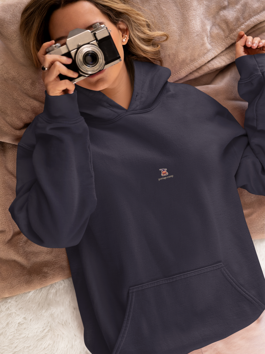 Cloud Ferry Midweight Hoodie in Classic Navy, worn by a woman in bed with a film camera, with the Cloud Ferry logo and portage.camp on the chest. Digital product mockup.