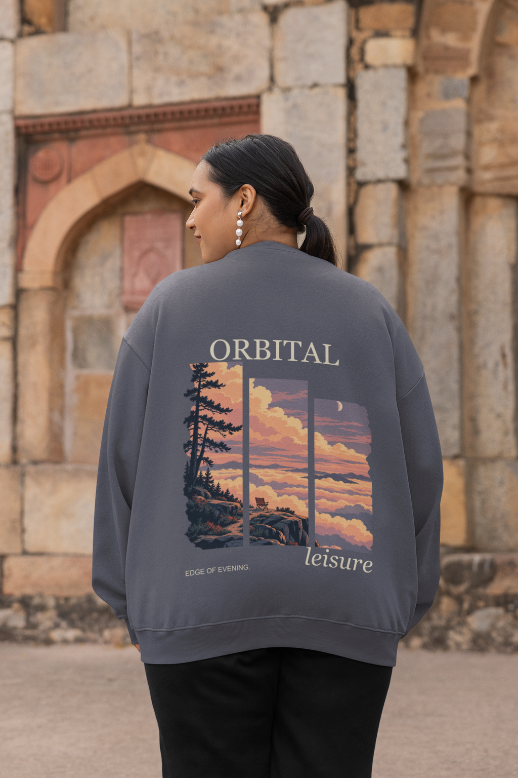 Edge of Evening Garment-Dyed Crewneck in Denim, worn by a woman seen from behind by an old stone building, showing the large print on the back. Digital product mockup.