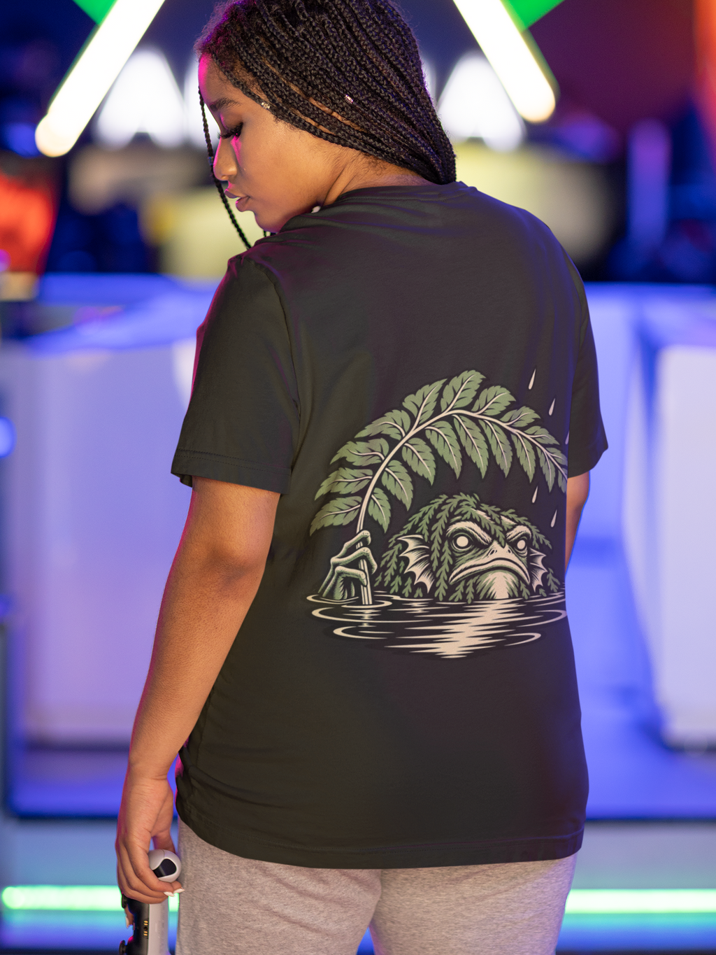 Rain Check Garment-Dyed Tee in Black, worn by a woman seen from behind under neon lights in a gaming arcade, showing the large print on the back. Digital product mockup.
