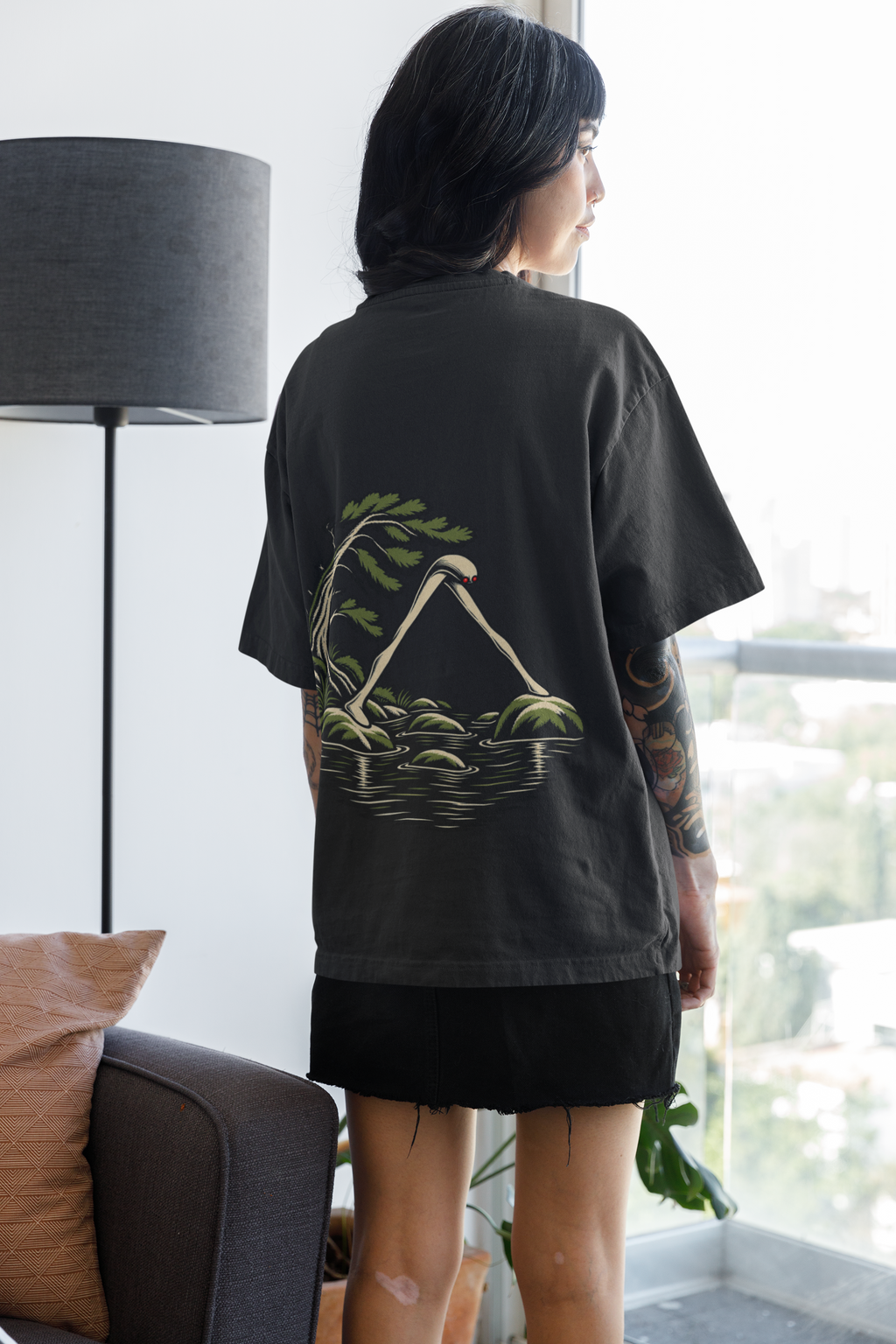 Small Hours Garment-Dyed Tee in Black, worn by a woman seen from behind at home by a bright window, showing the large print on the back. Digital product mockup.