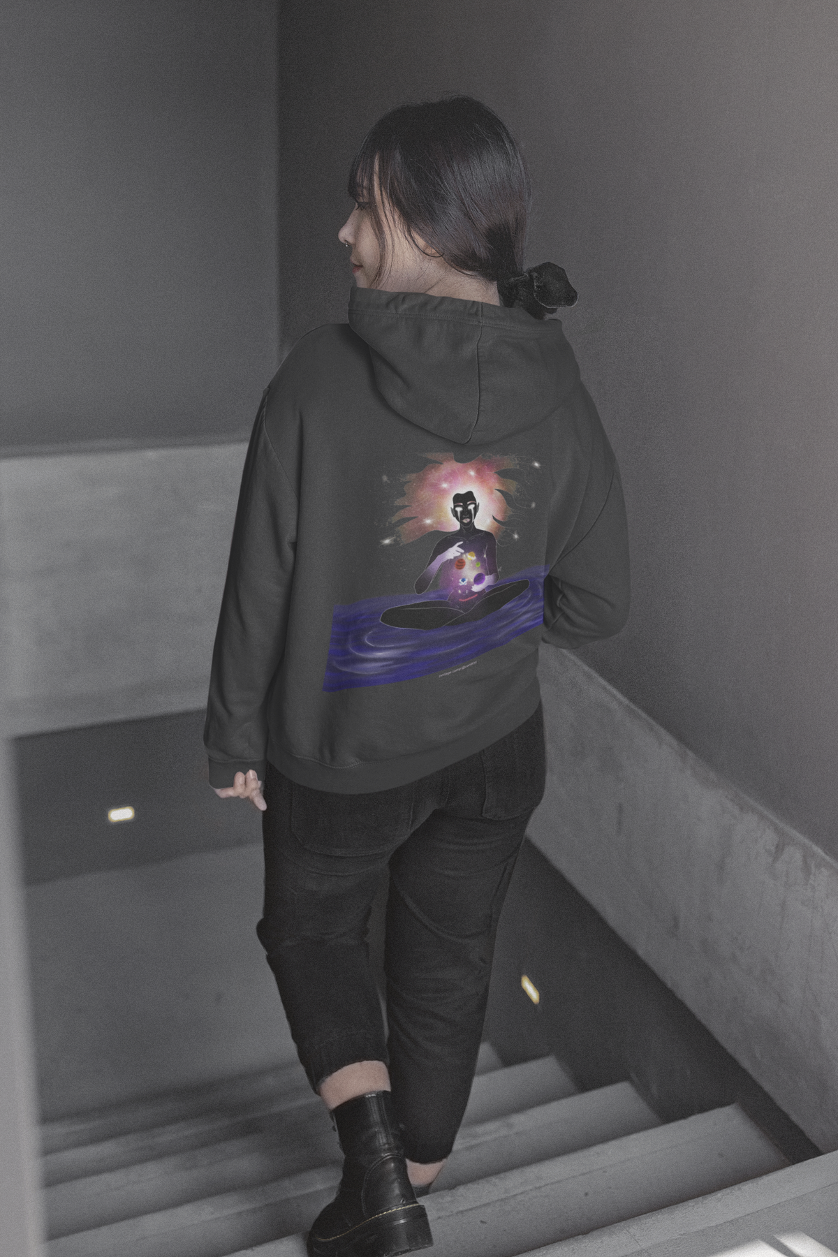 Orbit Keeper Heavyweight Hoodie in Black, worn by a woman seen from behind walking down a staircase, showing the large print on the back. Digital product mockup.