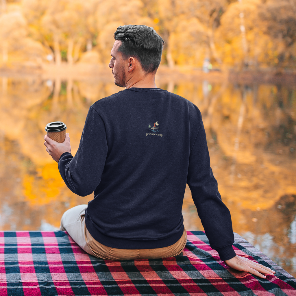 Quiet Hours Garment-Dyed Crewneck (Front Print) in True Navy, worn by a man seen from behind seated by a lake in autumn, showing the portage.camp emblem at the upper back. Digital product mockup.