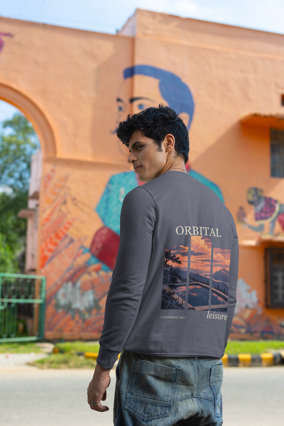 A Different Sky Garment-Dyed Crewneck in Denim, worn by a man seen from behind by a painted wall in the city, showing the large print on the back. Digital product mockup.