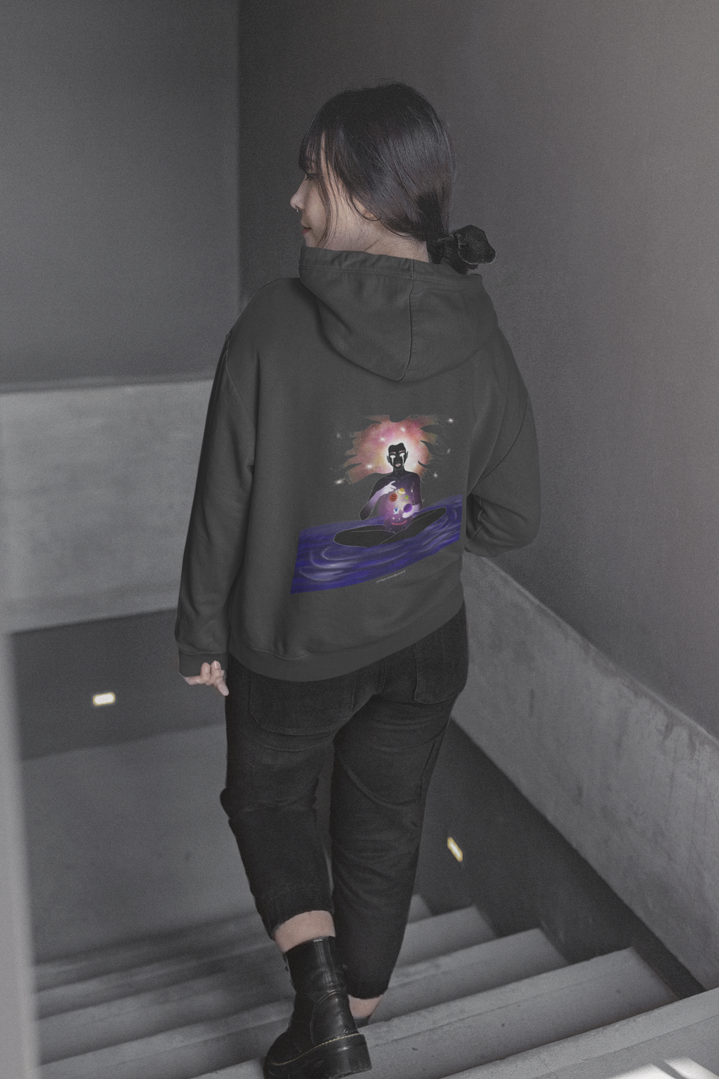 Orbit Keeper Heavyweight Hoodie in Black, worn by a woman seen from behind walking down a staircase, showing the large print on the back. Digital product mockup.