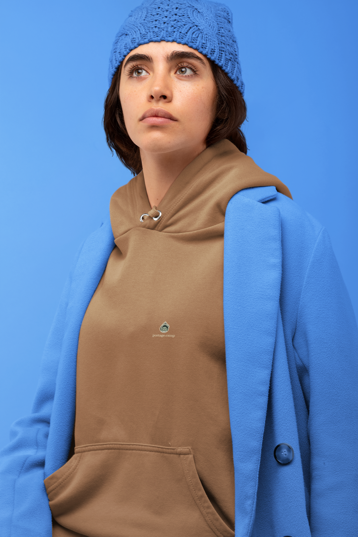 Rain Check Midweight Hoodie in Saddle, worn by a woman against a blue backdrop, with the Rain Check logo and portage.camp on the chest. Digital product mockup.