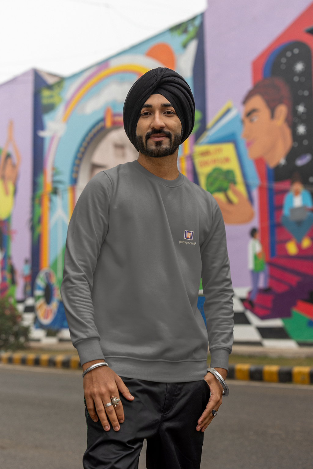 Open Late Garment-Dyed Crewneck in Pepper, worn by a man in a turban on a busy street, with the Open Late logo and portage.camp on the chest. Digital product mockup.