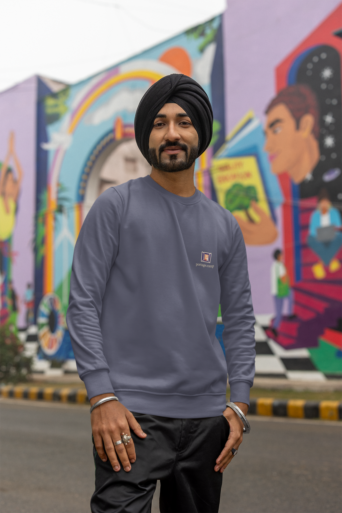 Open Late Garment-Dyed Crewneck in Denim, worn by a man in a turban on a busy street, with the Open Late logo and portage.camp on the chest. Digital product mockup.