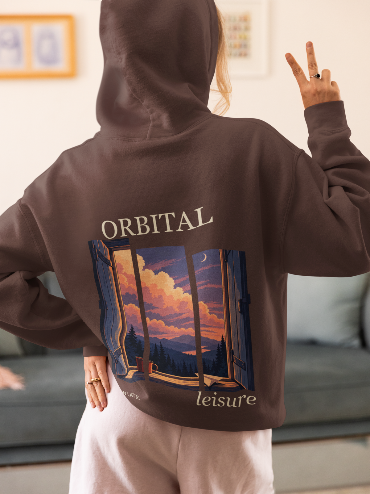 Cloud Ferry Heavyweight Hoodie in Brown, worn by a woman seen from behind at home, making a peace sign, showing the large print on the back. Digital product mockup.