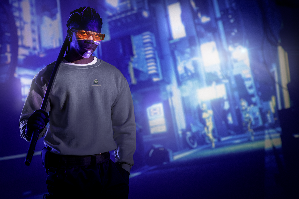 Rain Check Garment-Dyed Crewneck in Denim, worn by a man under neon city lights, with the Rain Check logo and portage.camp on the chest. Digital product mockup.