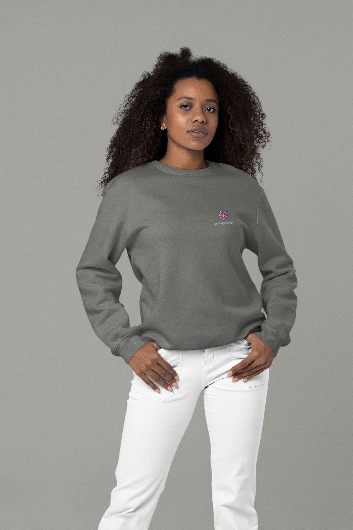 Heart Thief Garment-Dyed Crewneck in Pepper, worn by a woman in a studio with her hands in her pockets, with the Heart Thief logo and portage.camp on the chest. Digital product mockup.