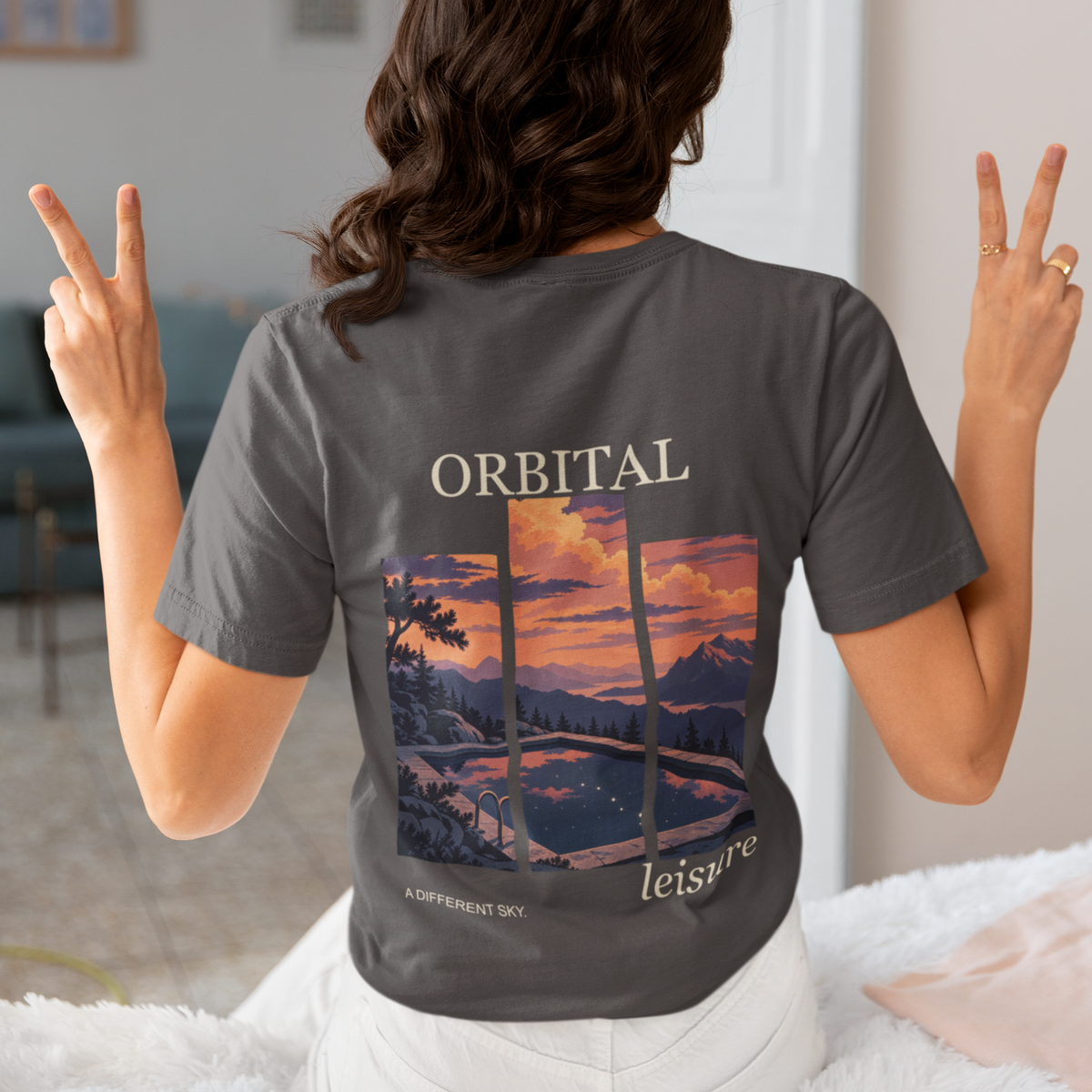 A Different Sky Garment-Dyed Tee in Graphite, worn by a woman seen from behind making peace signs, showing the large print on the back. Digital product mockup.