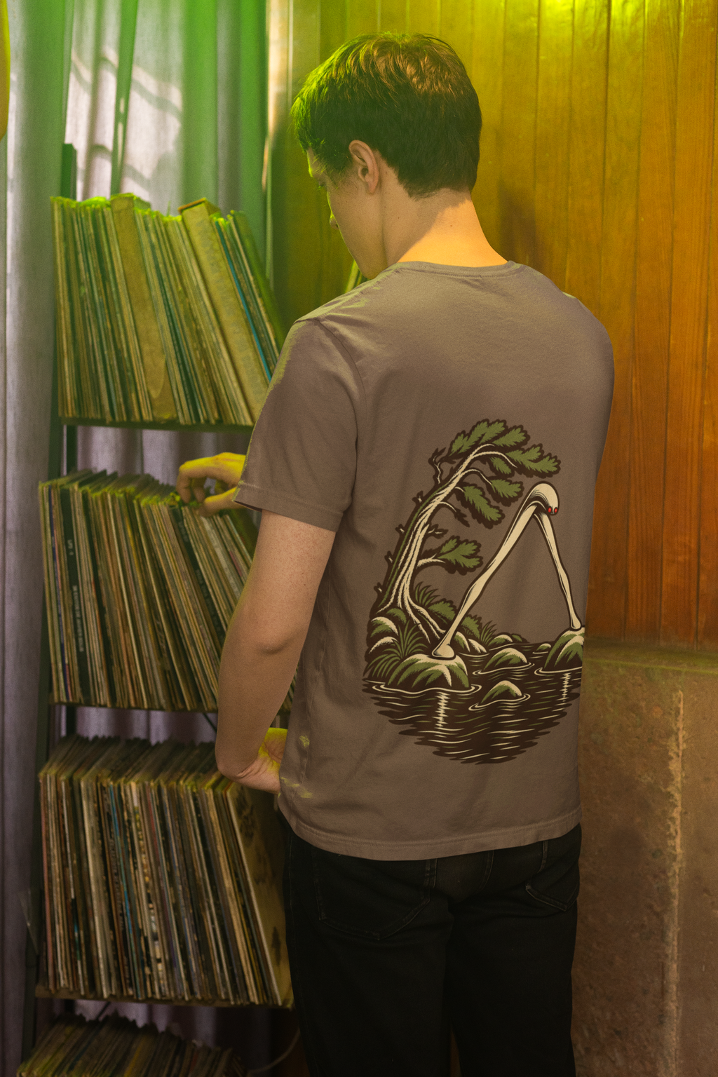 Small Hours Garment-Dyed Tee in Espresso, worn by a man seen from behind browsing a record shop, showing the large print on the back. Digital product mockup.