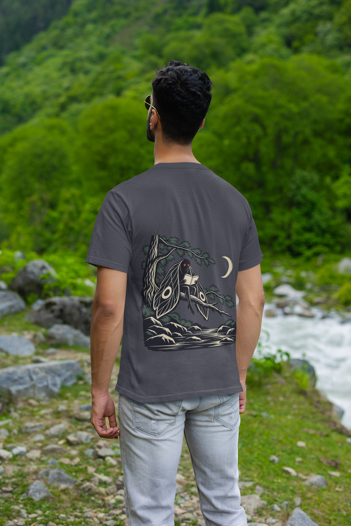 Night Library Garment-Dyed Tee in Navy, worn by a man seen from behind by a mountain river, showing the large print on the back. Digital product mockup.