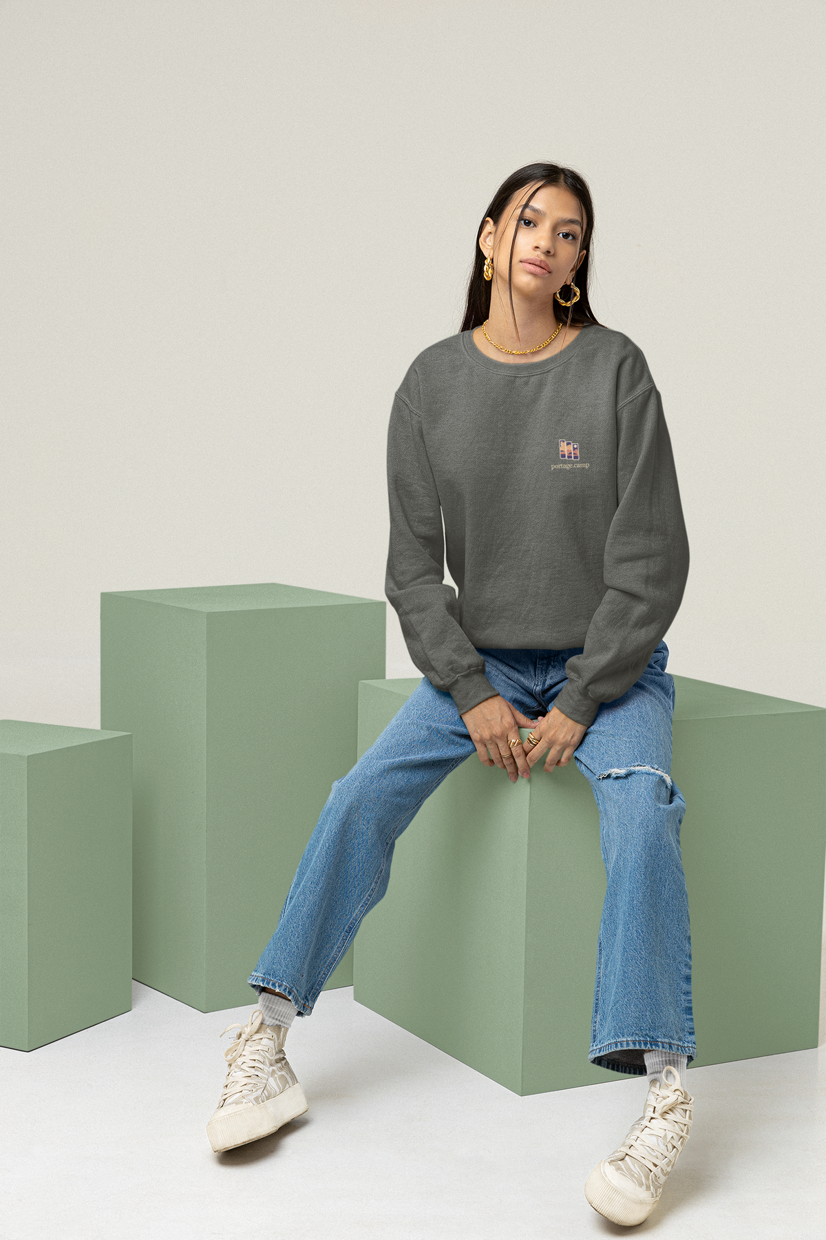Stay for the Sky Garment-Dyed Crewneck in Pepper, worn by a woman seated on green studio blocks, with the Stay for the Sky logo and portage.camp on the chest. Digital product mockup.