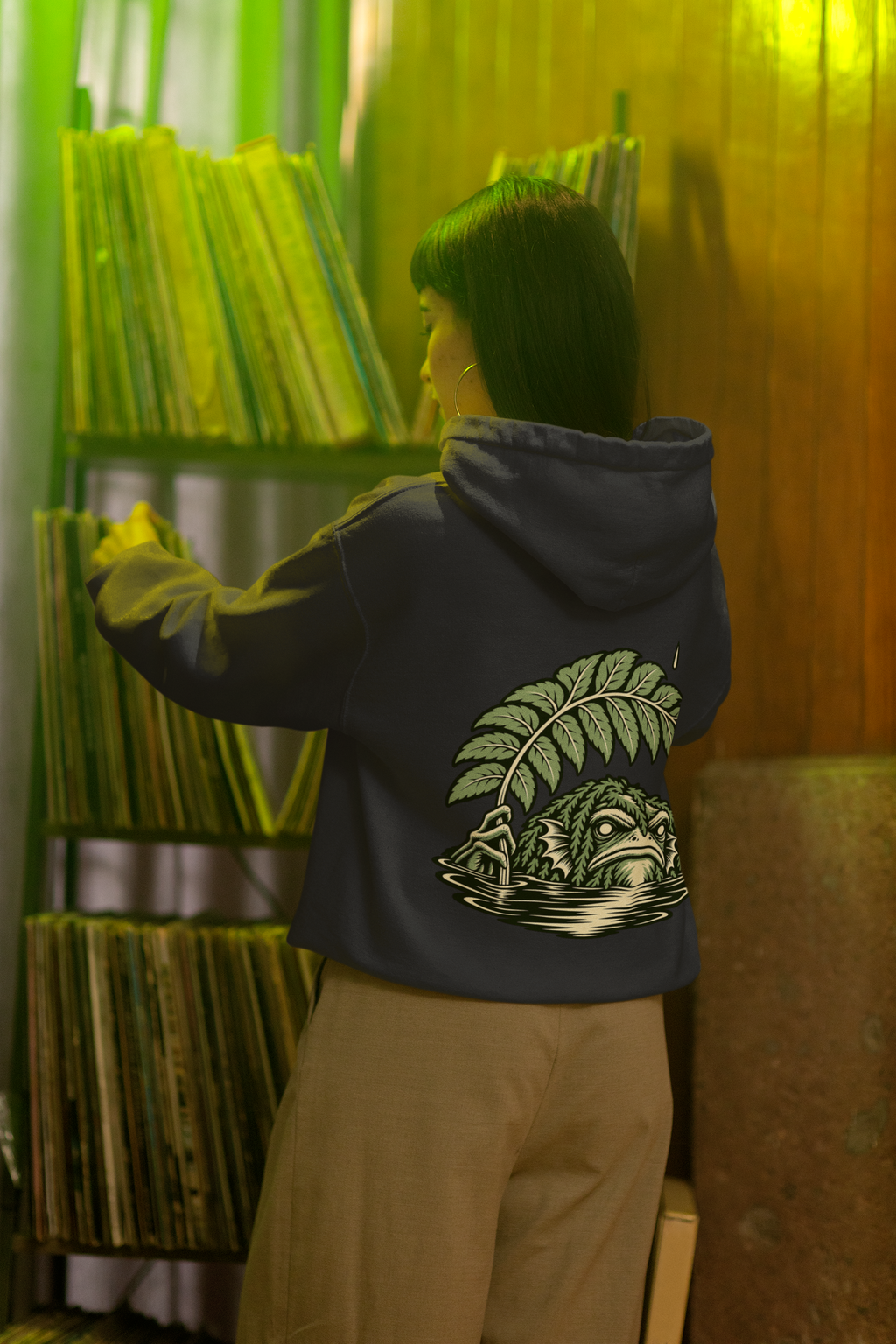 Seen Briefly Heavyweight Hoodie in Black, worn by a woman seen from behind in a vintage record shop, showing the large print on the back. Digital product mockup.