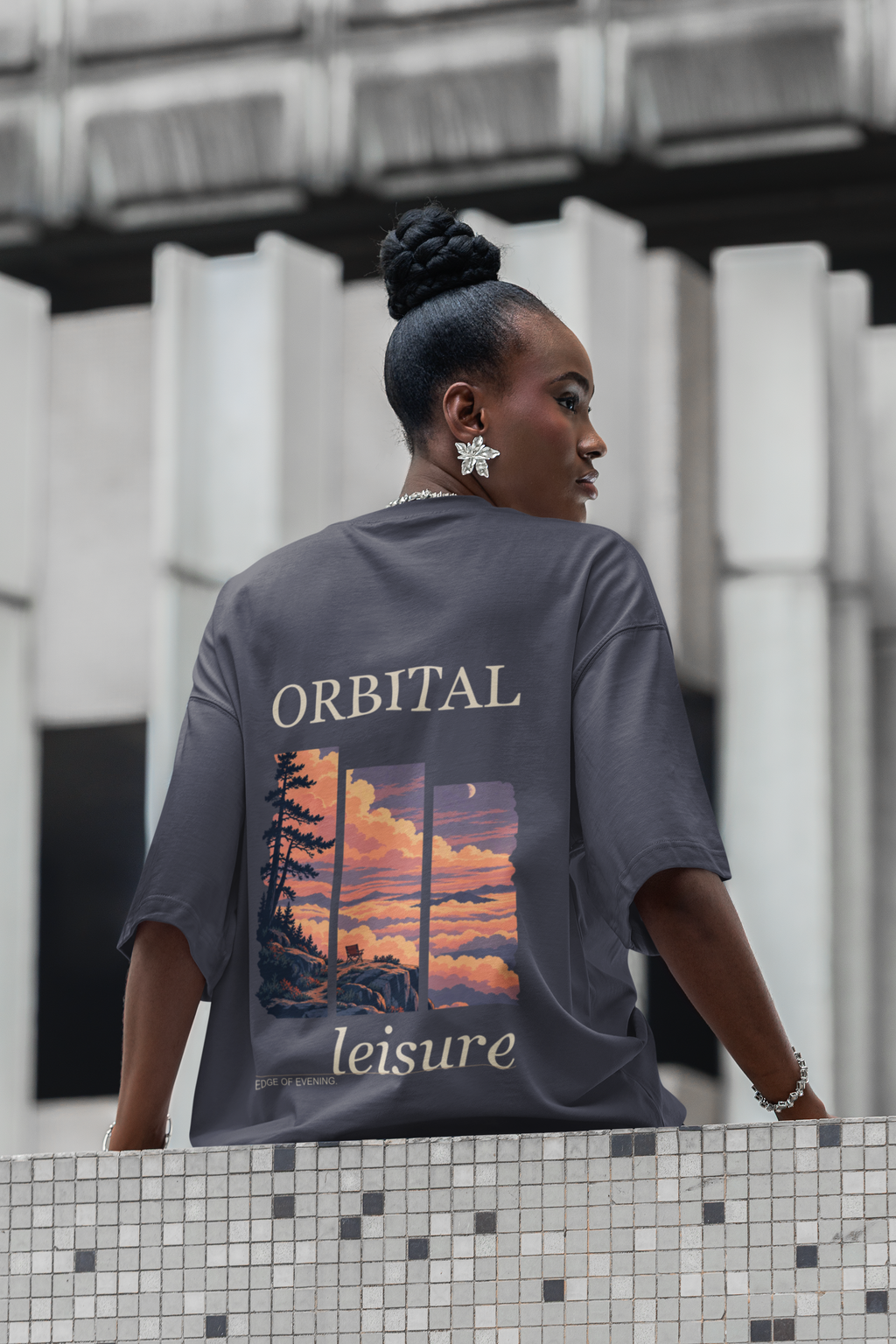 Edge of Evening Garment-Dyed Tee in Navy, worn by a woman seen from behind on a city street lined with columns, showing the large print on the back. Digital product mockup.