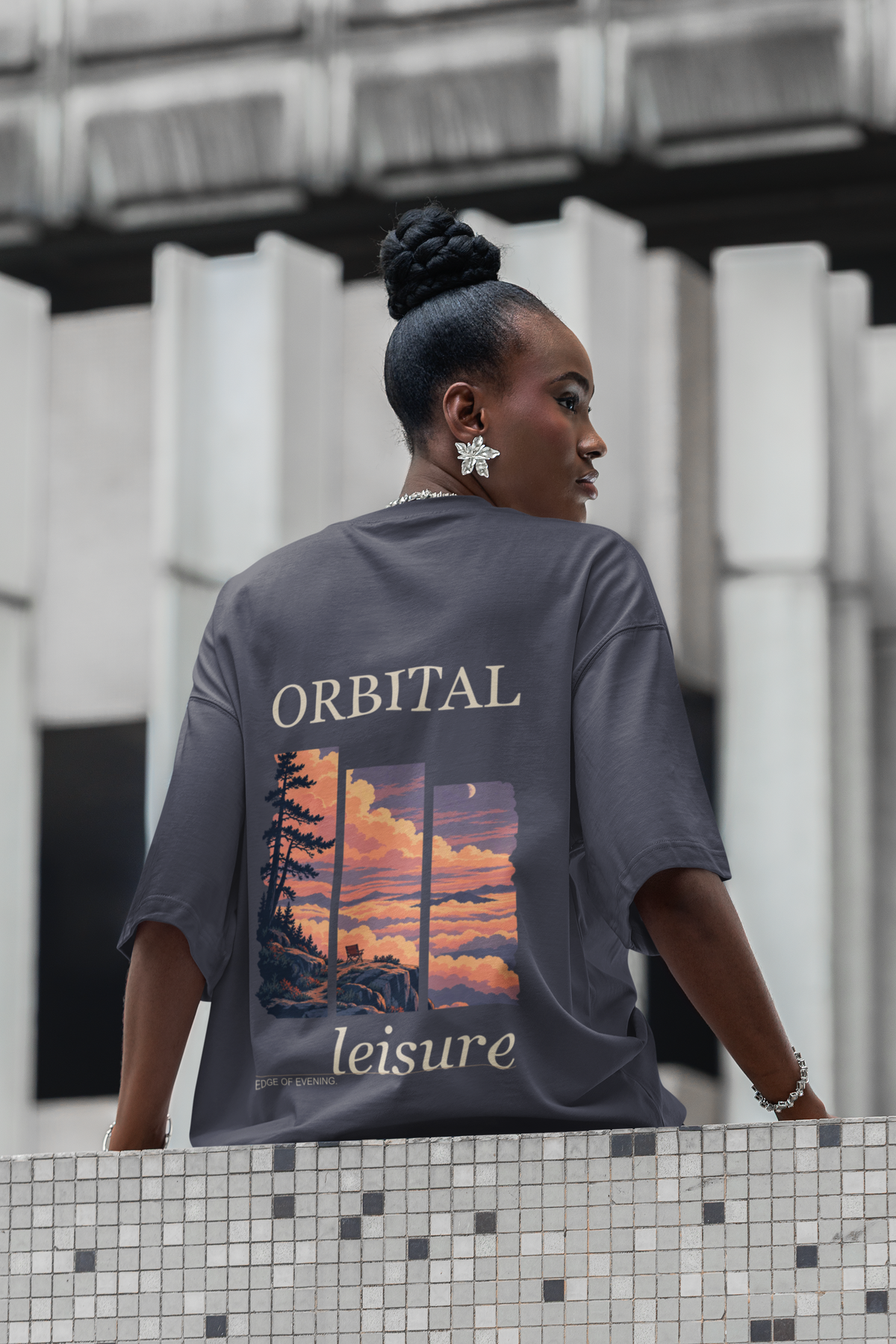 Edge of Evening Garment-Dyed Tee in Navy, worn by a woman seen from behind on a city street lined with columns, showing the large print on the back. Digital product mockup.