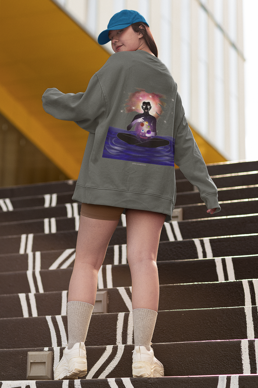 Orbit Keeper Garment-Dyed Crewneck in Pepper, worn by a woman seen from behind on outdoor stairs, showing the large print on the back. Digital product mockup.