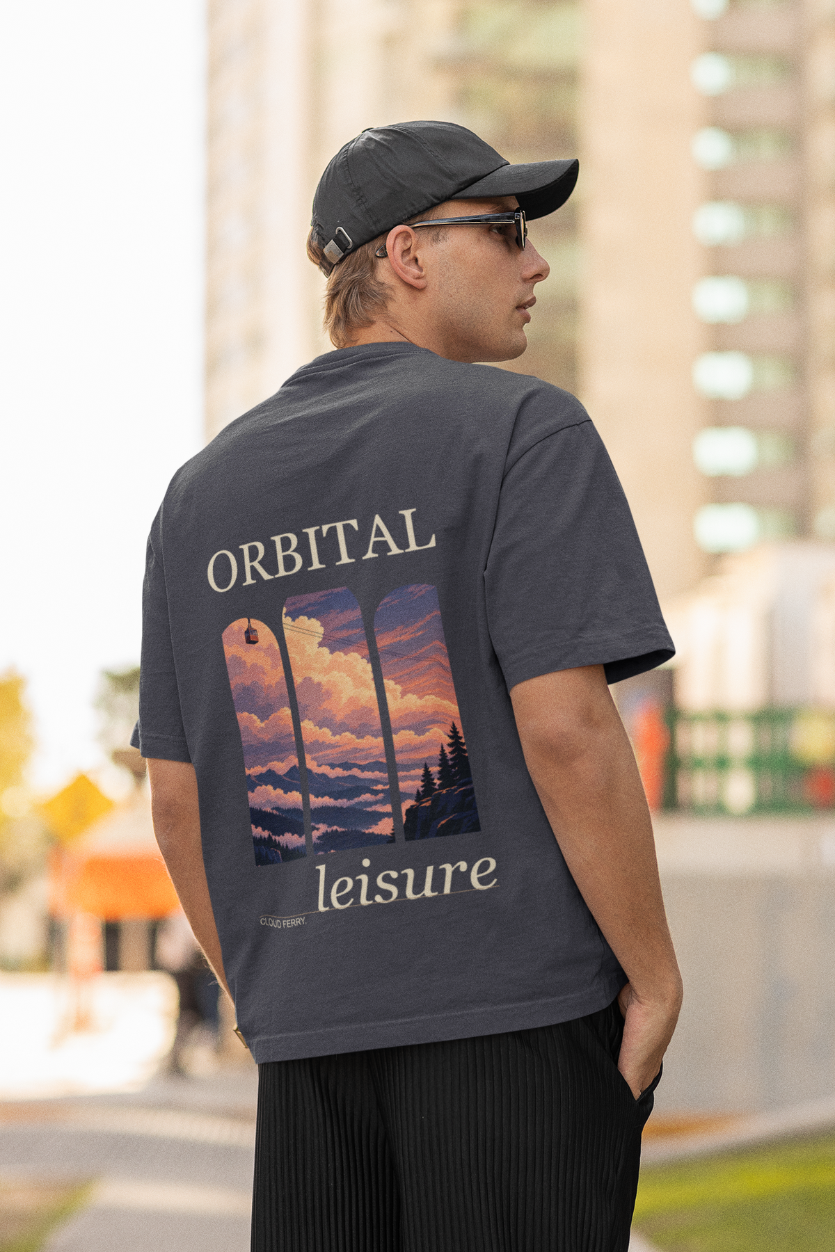 Cloud Ferry Garment-Dyed Tee in Navy, worn by a man seen from behind in a cap and sunglasses on a city street, showing the large print on the back. Digital product mockup.