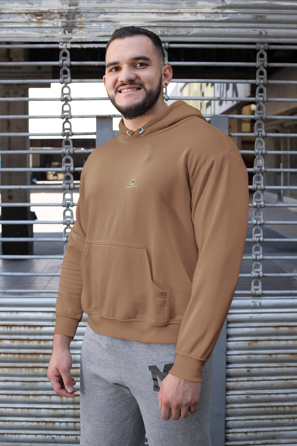 Rain Check Midweight Hoodie in Saddle, worn by a man against a metal fence, with the Rain Check logo and portage.camp on the chest. Digital product mockup.