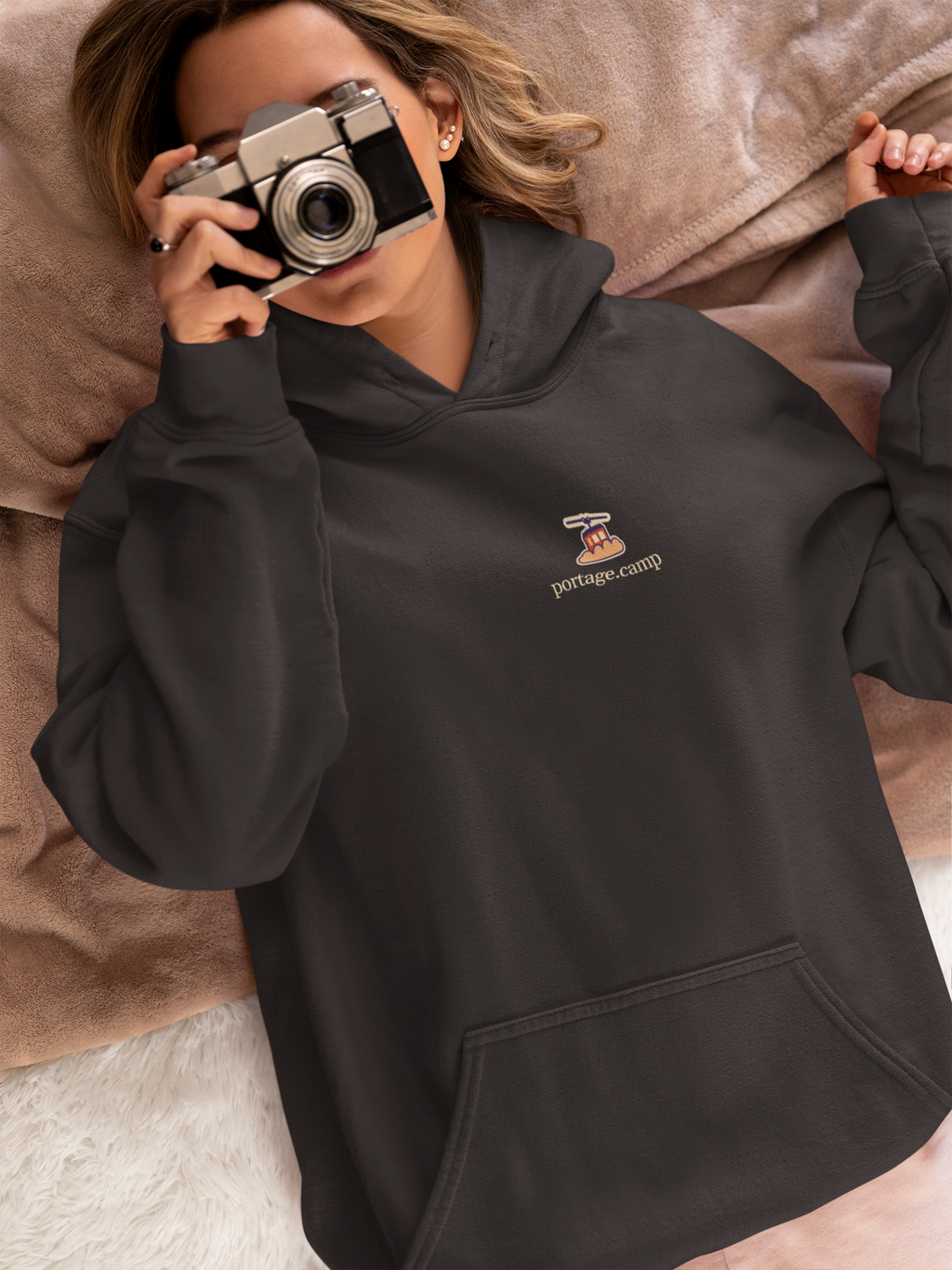 Cloud Ferry Heavyweight Hoodie in Black, worn by a woman in bed with a film camera, with the Cloud Ferry logo and portage.camp on the left chest. Digital product mockup.