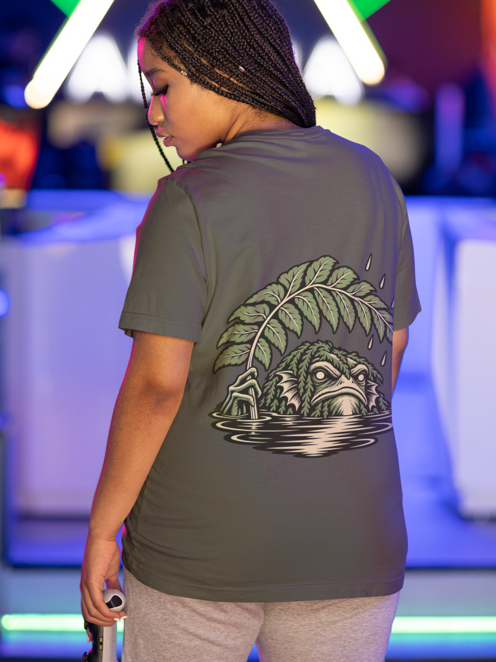 Rain Check Garment-Dyed Tee in Pepper, worn by a woman seen from behind under neon lights in a gaming arcade, showing the large print on the back. Digital product mockup.