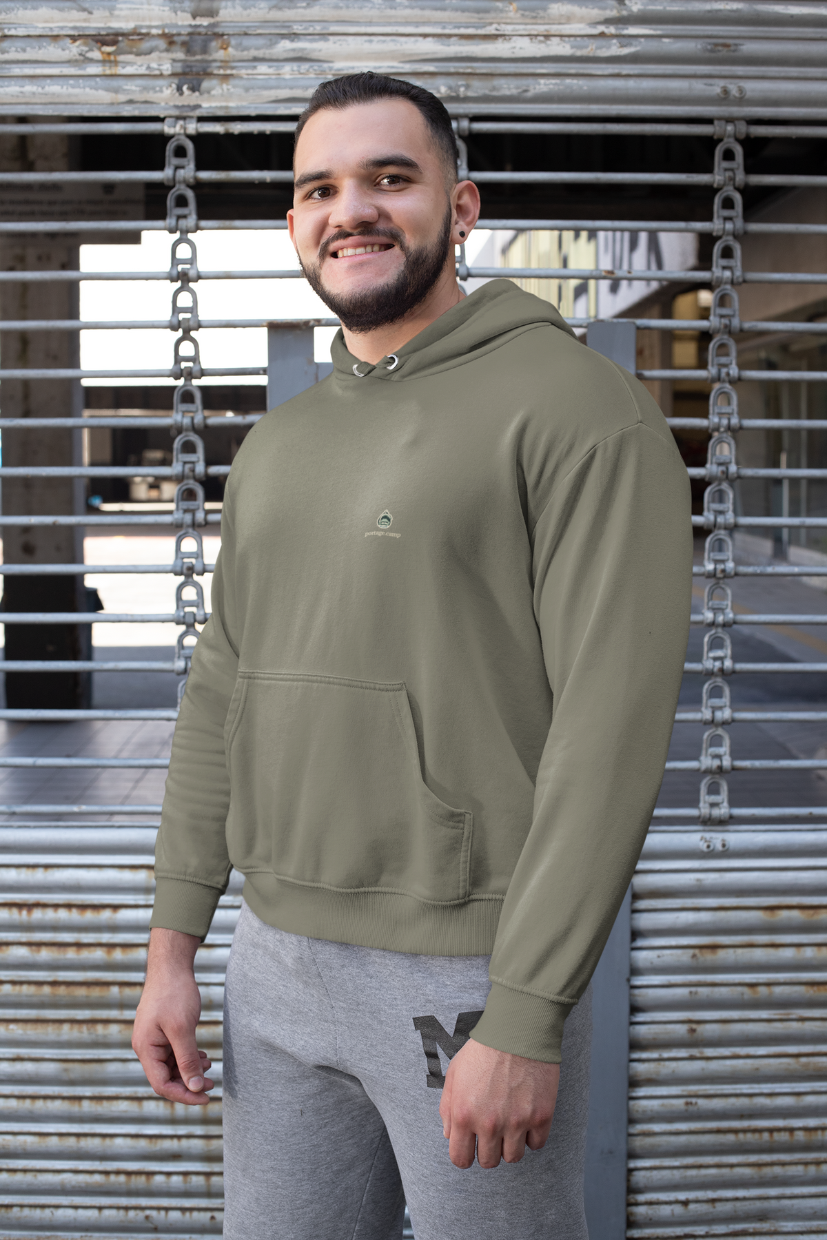 Rain Check Heavyweight Hoodie in Olive, worn by a man against a metal fence, with the Rain Check logo and portage.camp on the left chest. Digital product mockup.